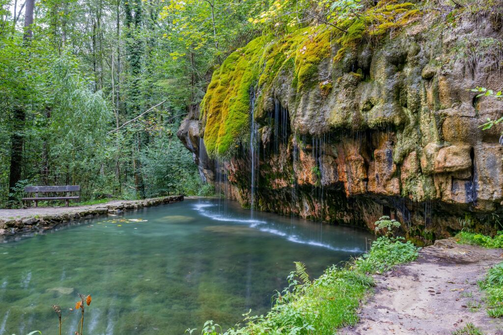 Wodospad Kallektuffquell na szlaku Mullerthal, źródło z krystaliczną wodą wapienną, pokryte mchem formacje skalne z piaskowca, spadająca woda, drzewa i brzeg w tle, Luksemburg